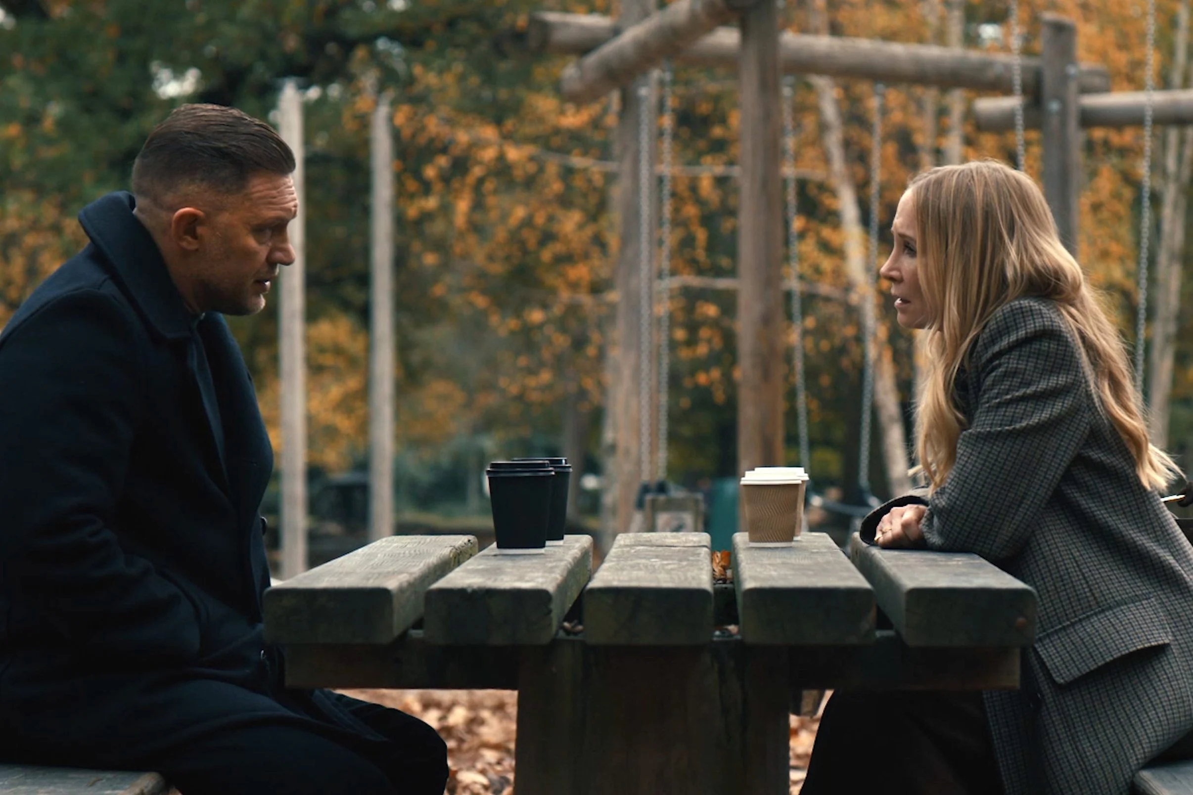 Tom Hardy and Joanne Froggatt sat outside in a park at a table, opposite one another in MobLand.